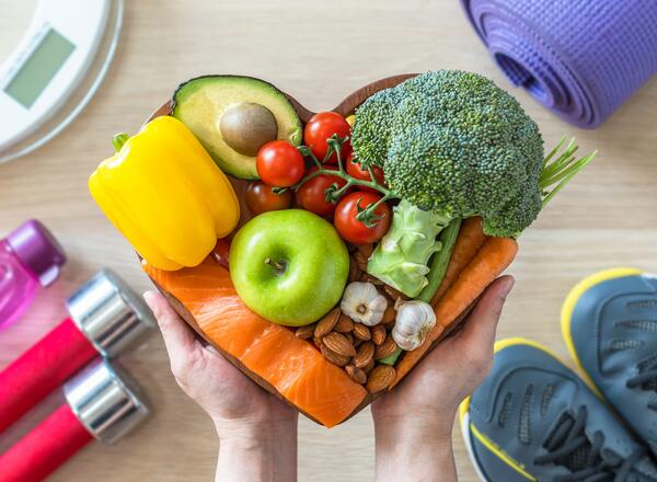 Hands holding a basket of fruits and vegetables shaped like a heart with fitness equipment on the floor